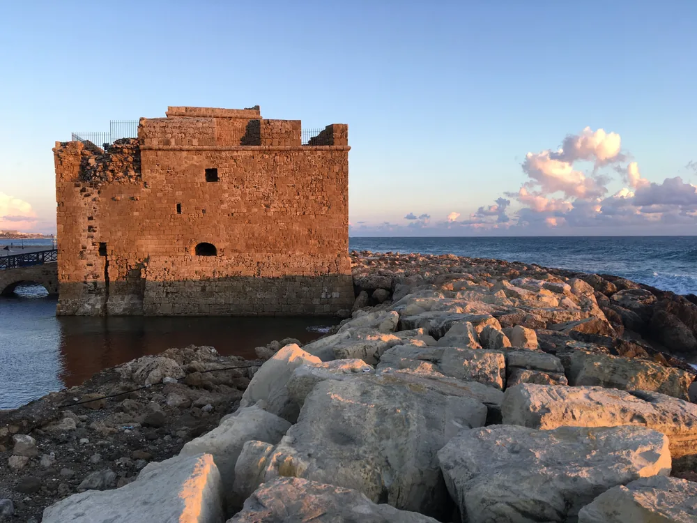 Paphos Castle, eine quadratische Steinfestung direkt am Wasser, die bei Sonnenuntergang neben einem felsigen Wellenbrecher golden leuchtet