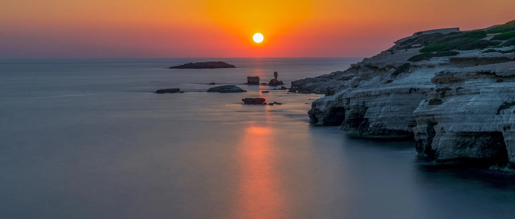 Eine helle Kalksteinküste und eine kleine Insel auf ruhigem Wasser, während die Sonne am Horizont untergeht