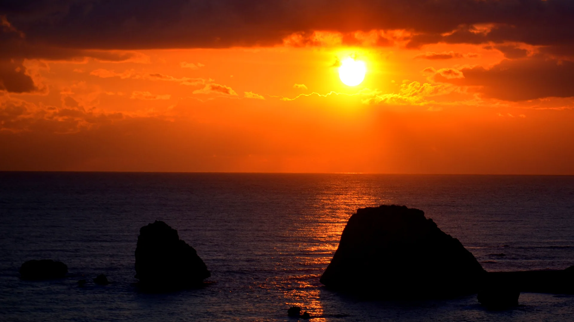 Die Sonne geht durch einen orangefarbenen Himmel über ein ruhiges Meer unter, dunkle Felsen im Wasser zeichnen sich als Silhouetten ab