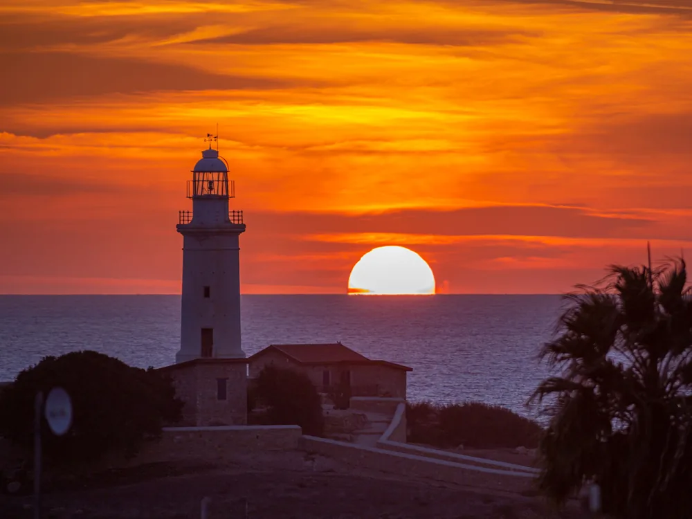 Ein weißer Leuchtturm, der sich vor einem tief orangefarbenen Himmel abzeichnet, während die Sonne das Meer berührt